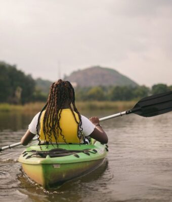 Person paddling a green kayak on a river near wooded hills