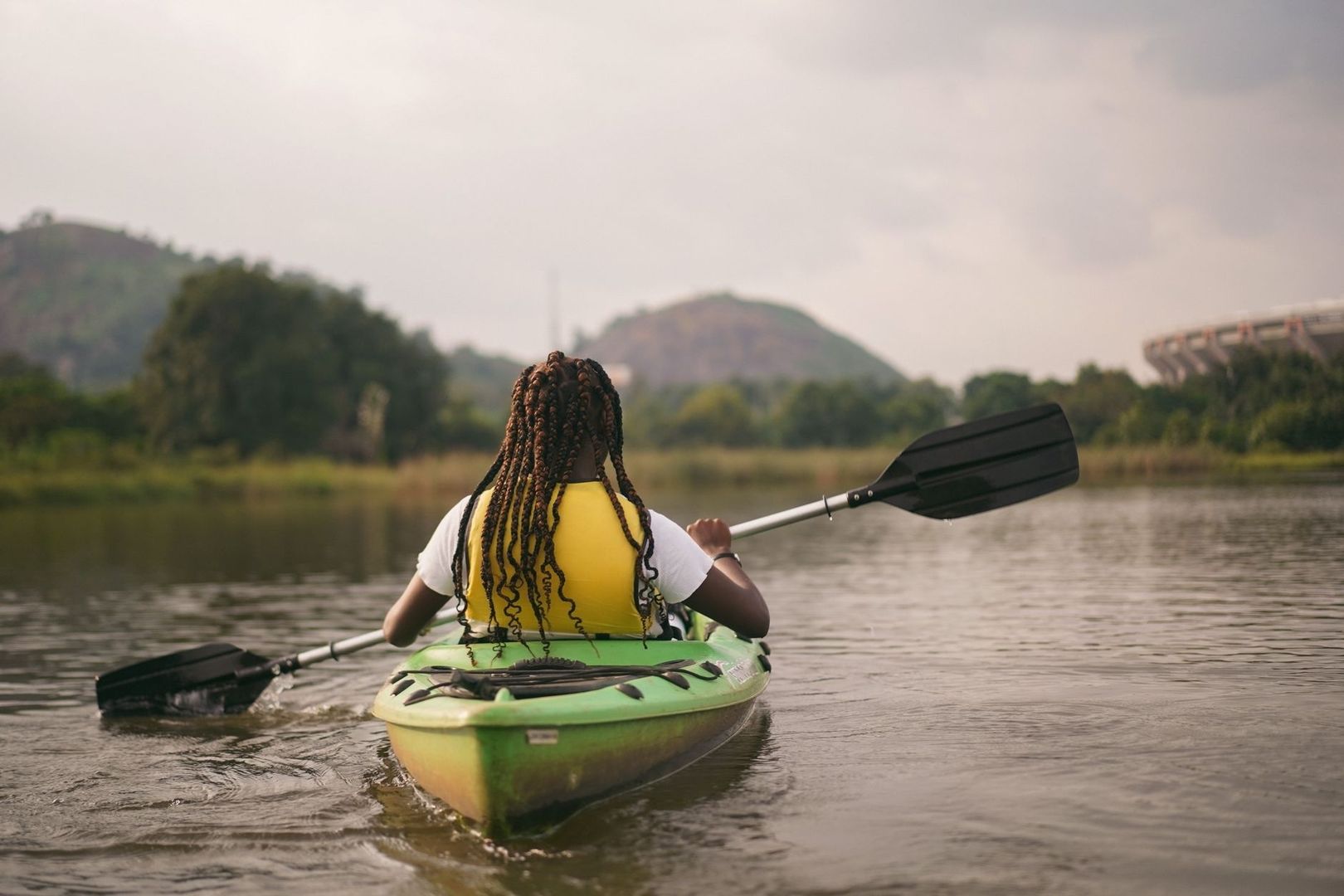 Person paddling a green kayak on a river near wooded hills